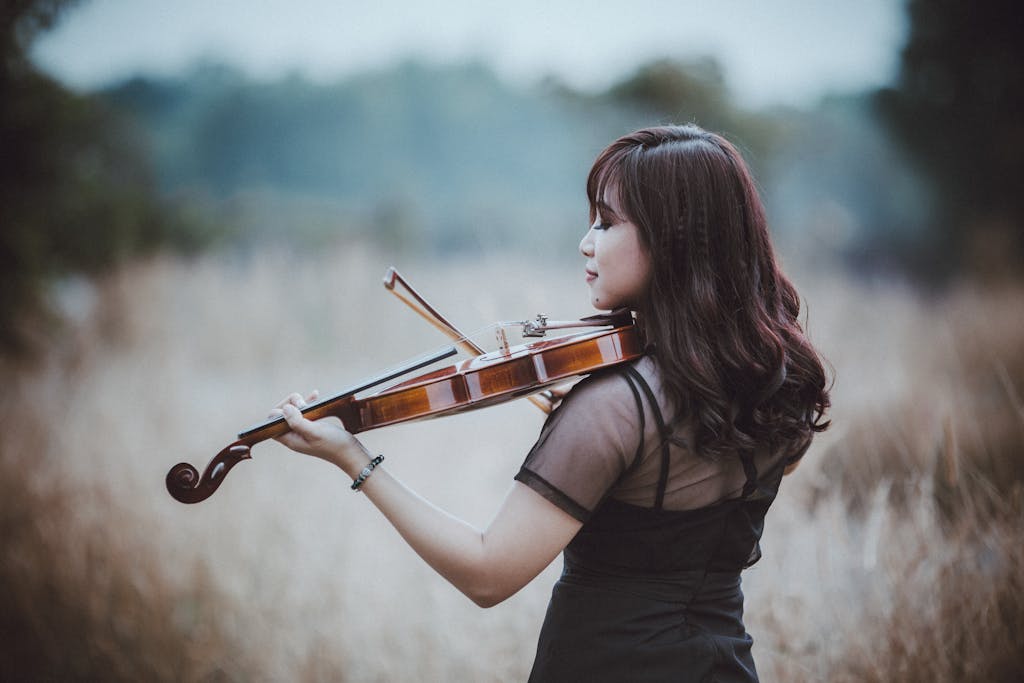 Back view of young ethnic female musician in elegant apparel with closed eyes playing violin on meadow